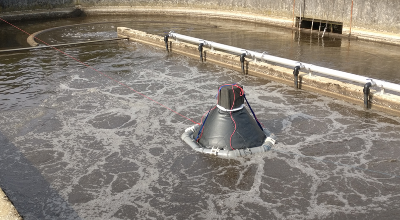 A photograph of our offgas hood. It shows a tank of a sewage treatment plant from above. The tank is filled with brown wastewater. A pyramid-shaped construction containing our measuring technology for offgas analysis floats on the water.