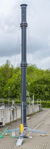 Photograph of our testing facility for determining the alpha value. It shows a 7 meter high, approximately 40 centimetre thick pipe. The pipe consists of several individual parts and stands on a steel tripod. The background shows a tank of a sewage treatment plant.
